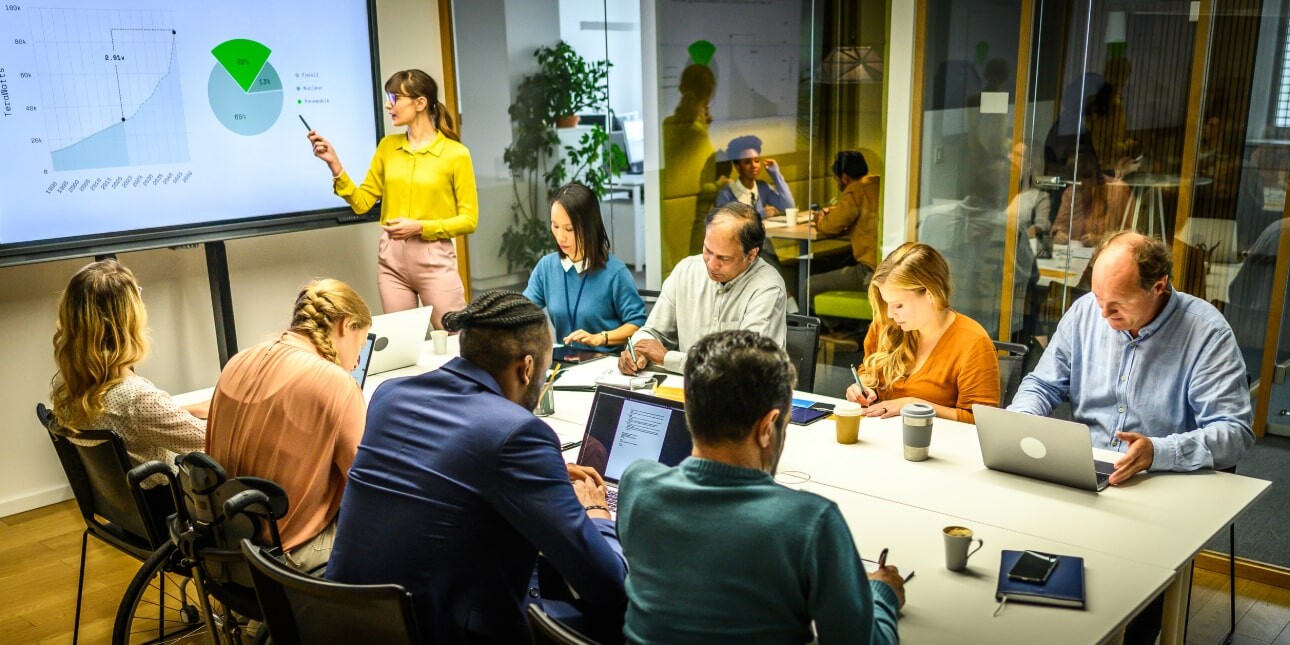 In a meeting room eight colleagues of different ages, ethnicity and gender are sat with laptops around a table. A woman dressed in yellow is stood pointing at a presentation on a screen.